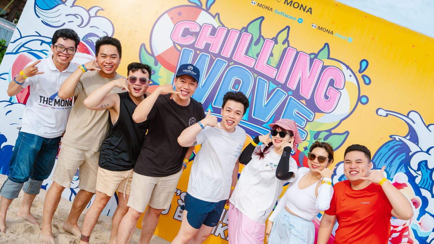 Nhân viên MONA tạo dáng trước phông team building Chilling Wave bên biển